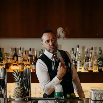 Barkeeper mit Weste und Fliege hantiert mit rauchendem Cocktail vor Spirituosenregal