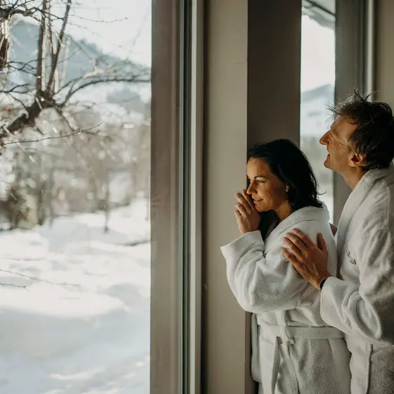 Paar in Bademänteln blickt aus Fenster auf verschneite Winterlandschaft