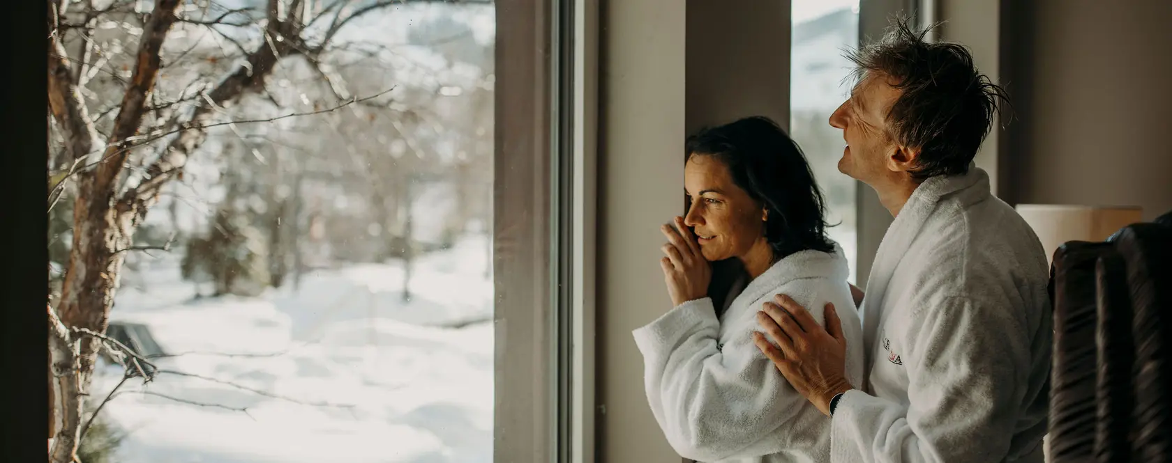Paar in Bademänteln blickt aus Fenster auf verschneite Winterlandschaft