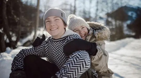Two laughing children hugging in the snow, wearing winter clothes and hats