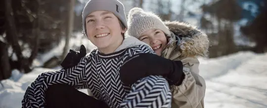 Zwei lachende Kinder umarmen sich im Schnee, tragen Winterkleidung und Mützen
