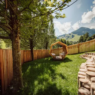 Garten mit Holzzaun, Baumstämmen und einer sechseckigen Holzkonstruktion vor Bergkulisse