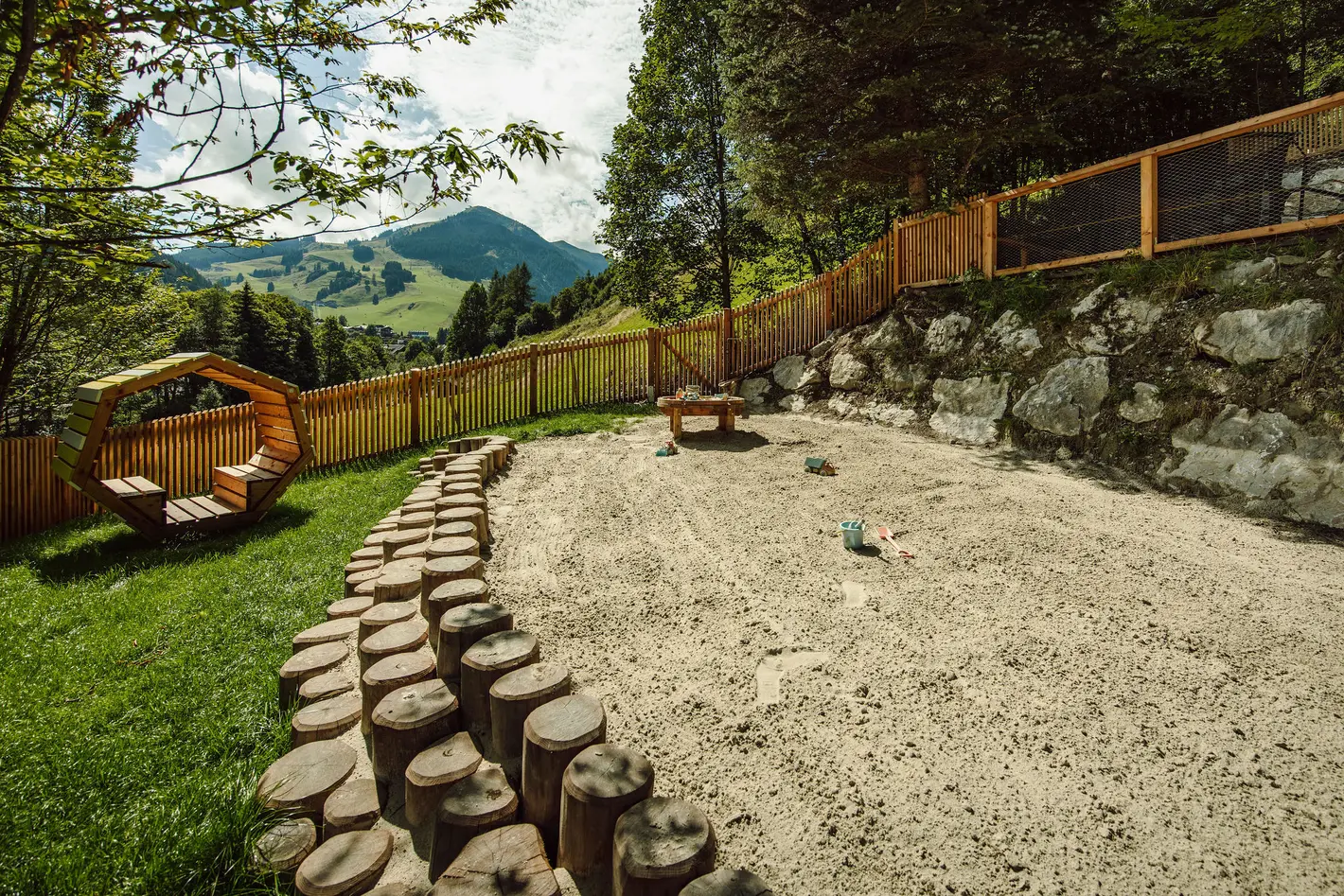 Spielplatz mit Holzzaun, Baumstämmen als Weg und Bergpanorama im Hintergrund