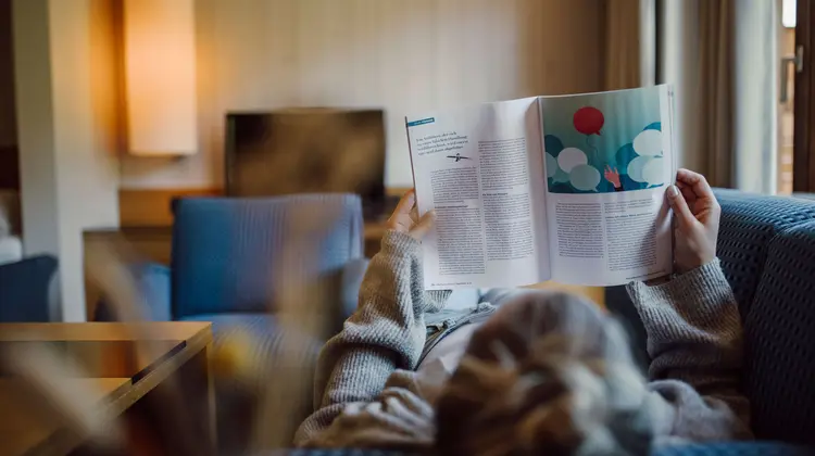 Person liest Zeitschrift auf Couch, TV im Hintergrund