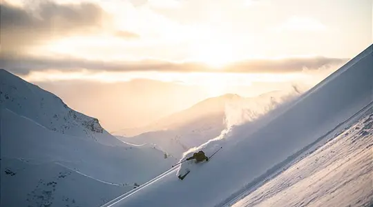 Skier skiing down a steep slope, cloud of dust, mountain panorama in the golden evening light